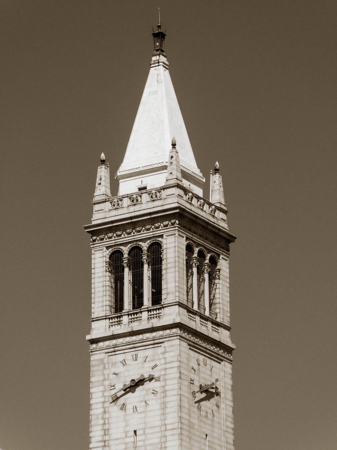 the campanile at uc berkeley in sepia, clock faces and white spire against a clear sky