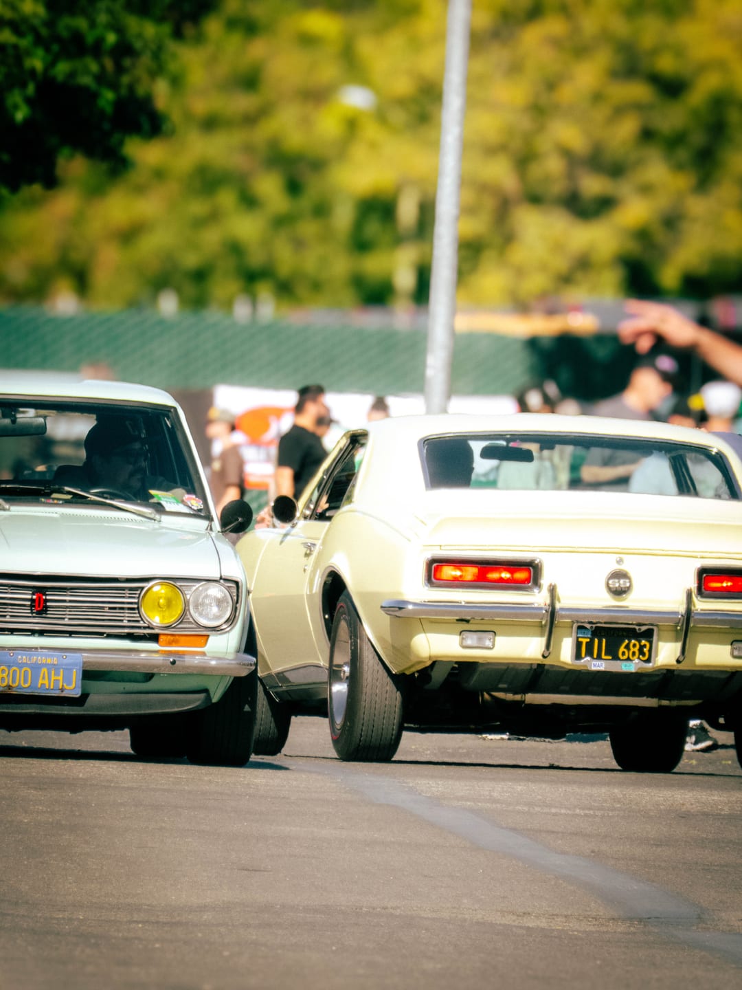 mint green datsun nose to tail with a pale yellow camaro ss at a car show, crowd behind