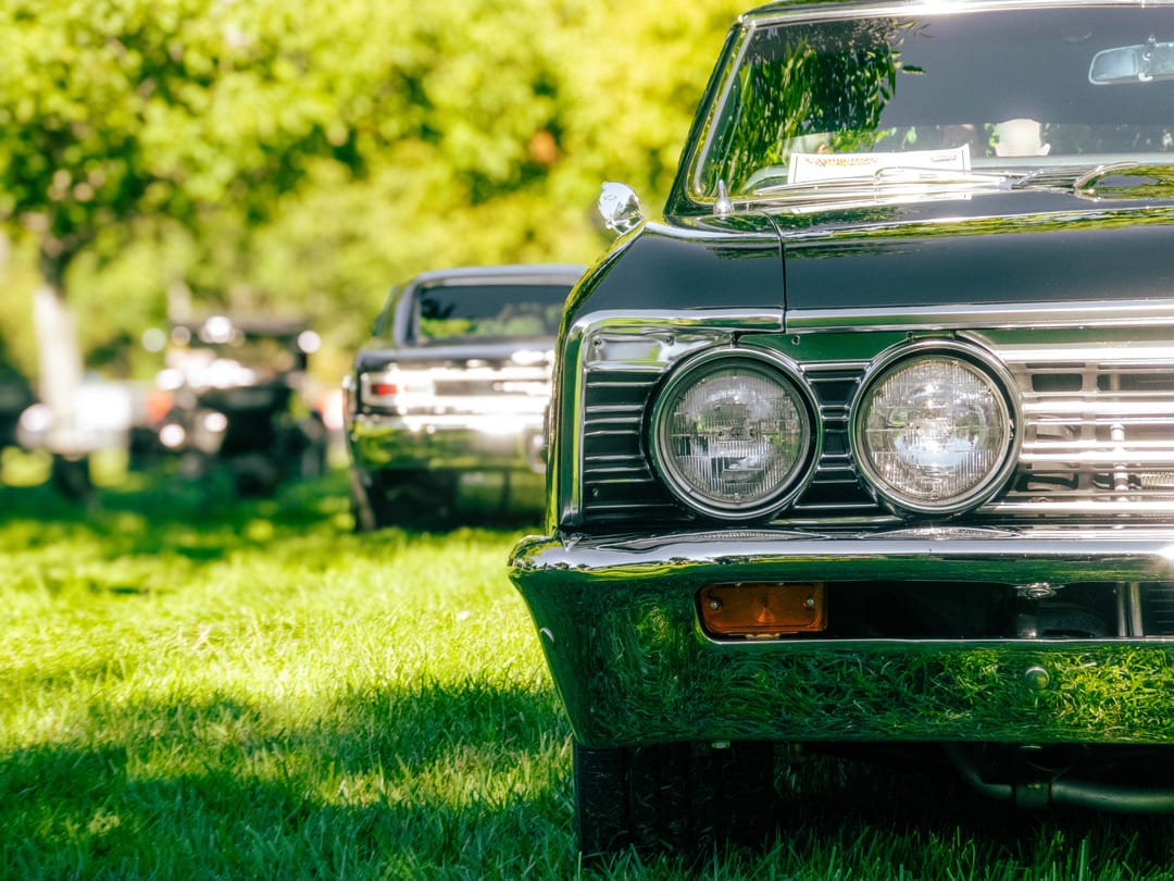 quad headlights and chrome bumper of a dark muscle car on a lawn, another classic parked behind