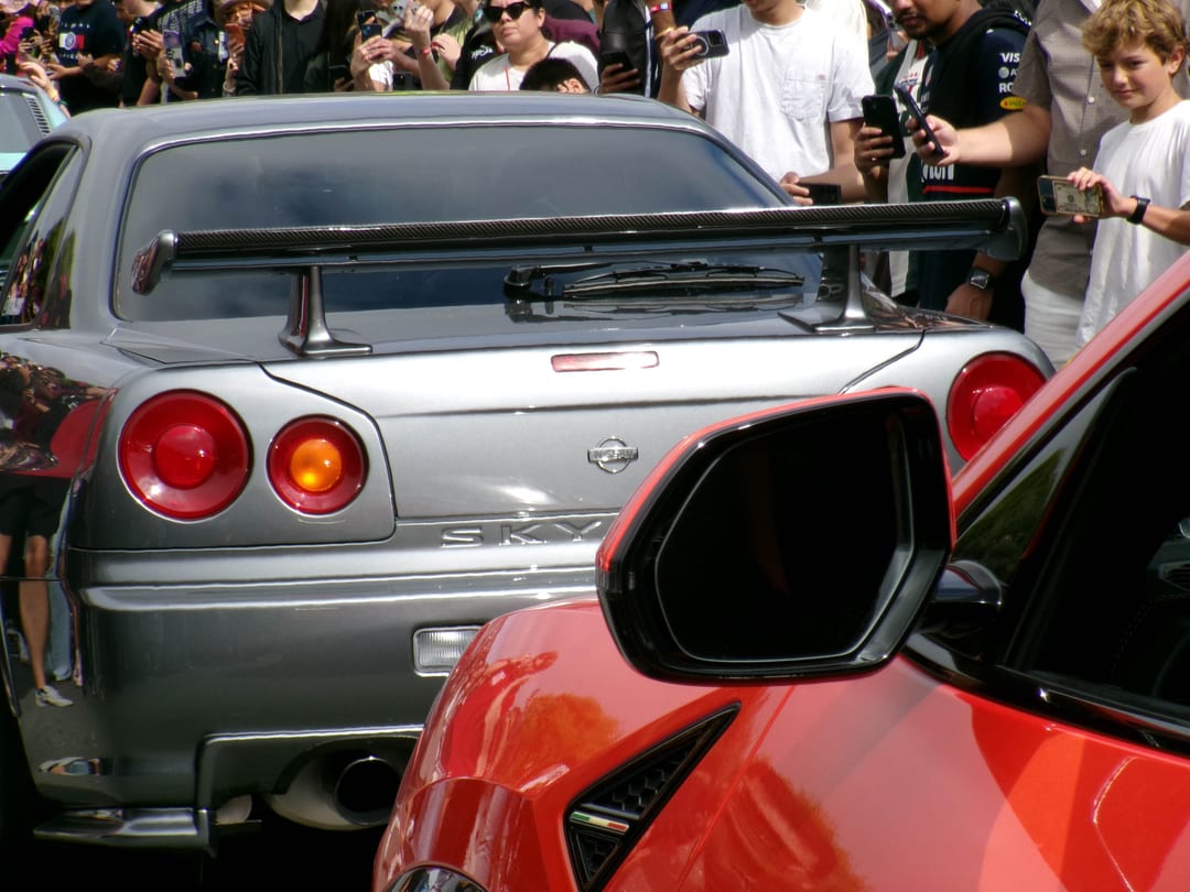 rear of a silver nissan skyline gt-r r34 with a carbon wing, red car mirror in the foreground