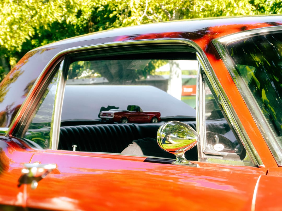 through the side window of a red classic car: a toy red mustang on the dash and a round chrome mirror
