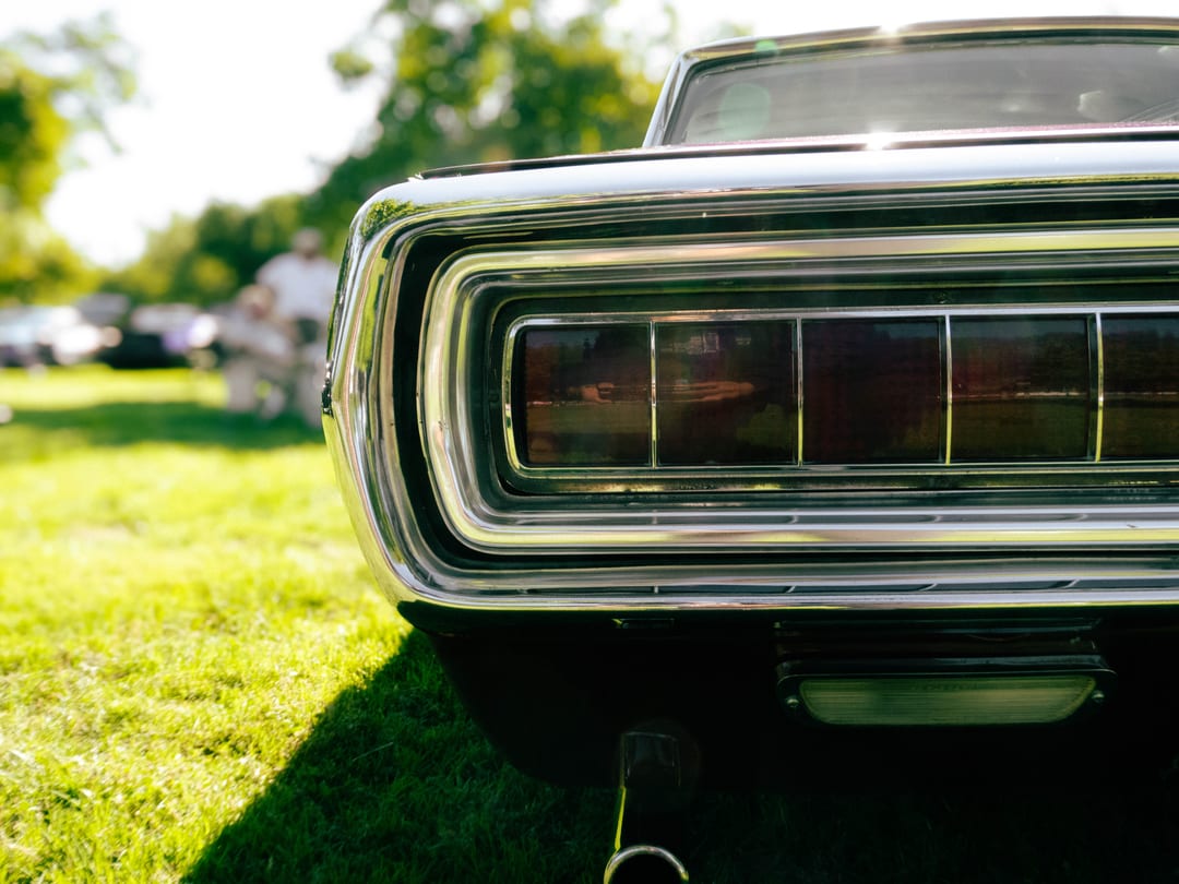 wide chrome-framed tail light of a late-sixties car parked on grass, people blurred behind