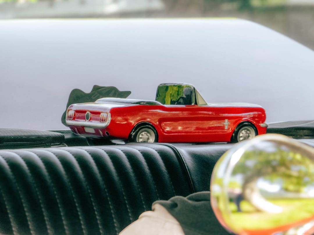 a red toy mustang convertible sitting on a black dashboard, chrome side mirror in the corner